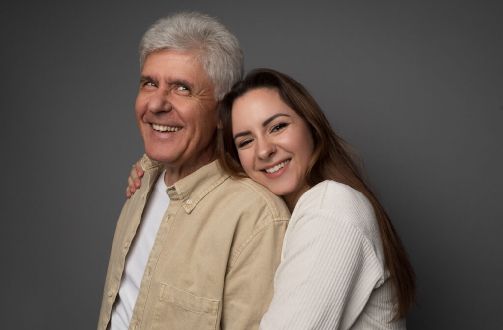 A senior man grinning as his daughter embraces his shoulder with a warm smile