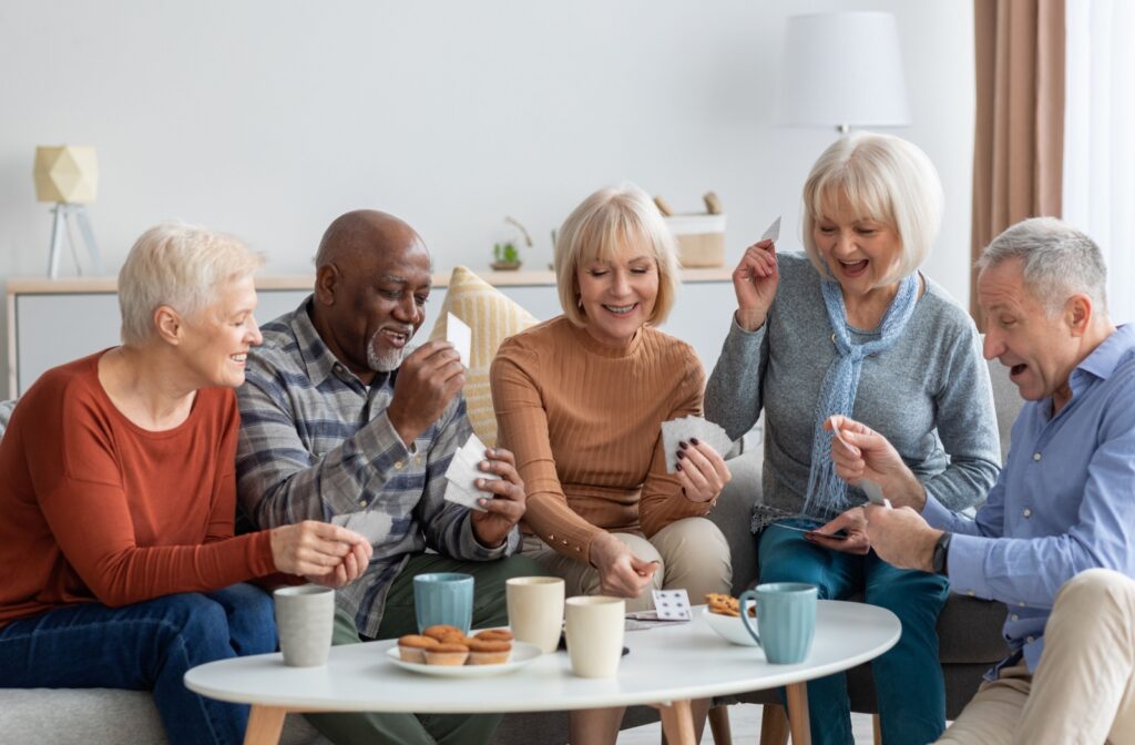 A group of older adults at a senior living community sit around a table laughing and playing cards together