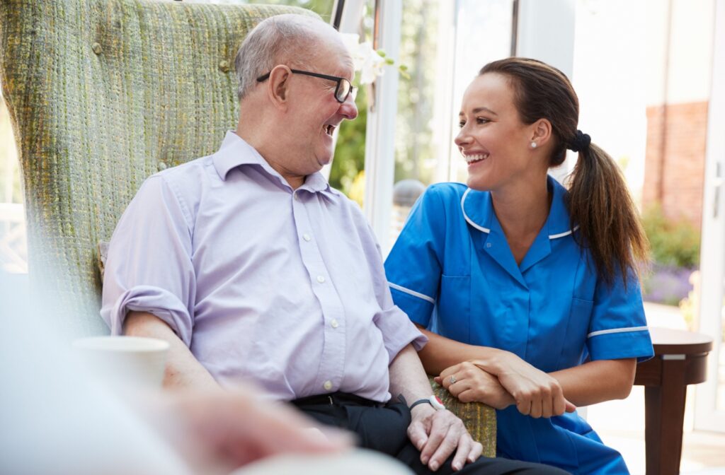 An older adult man in a memory care community sitting on a chair smiling and conversing with a nurse.