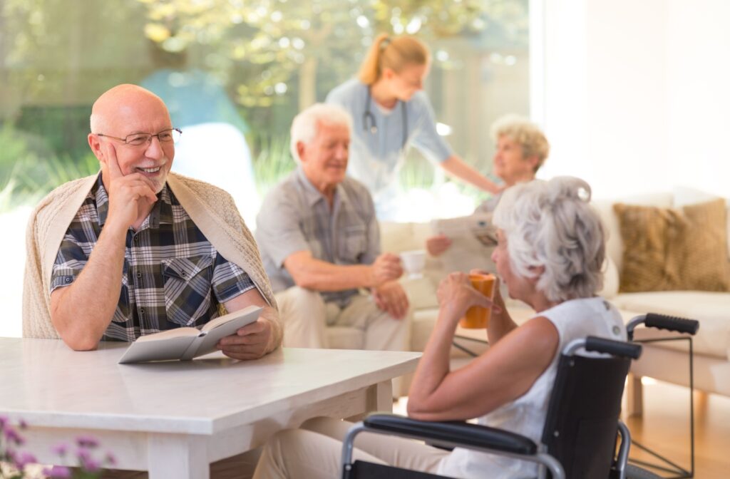 Seniors together in a memory care community. Some sitting at a table enjoying some tea and reading a book, while others are in the background chatting with a nurse.