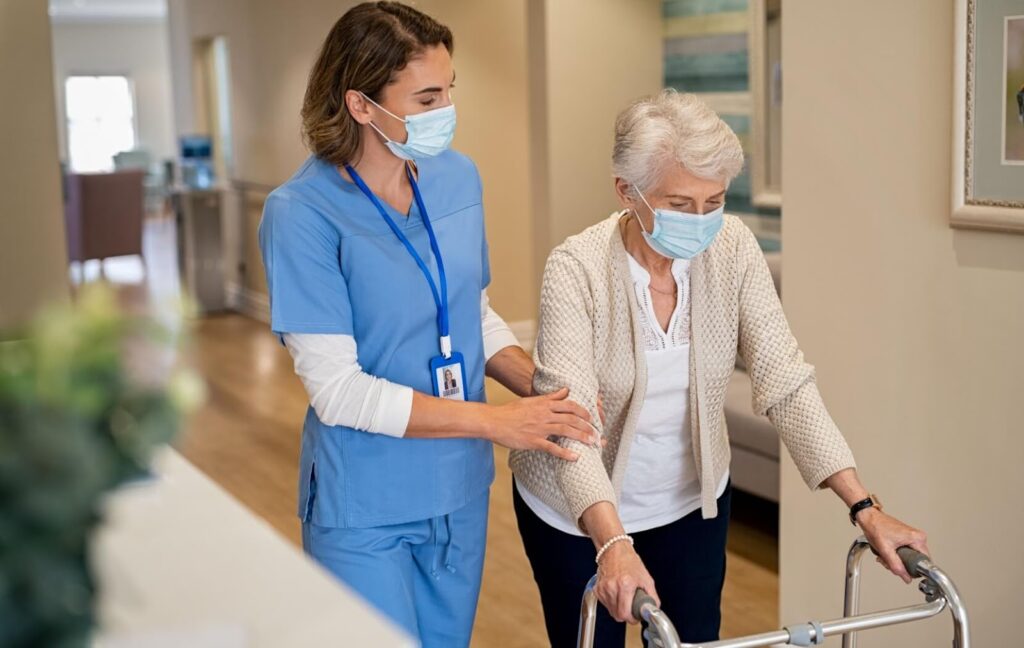 A nurse senior a resident walk down a hallway. The nurse hold's the senior's arm and the senior is using a walker.