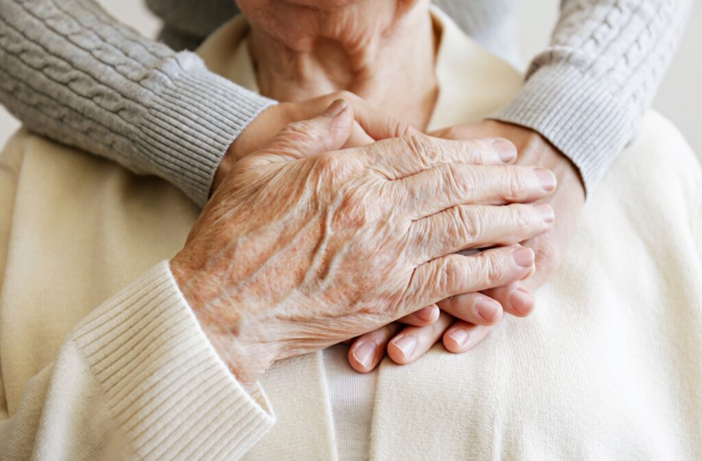 An adult child embraces their senior parent. Their senior parent reaches up to grasp their child's hands.