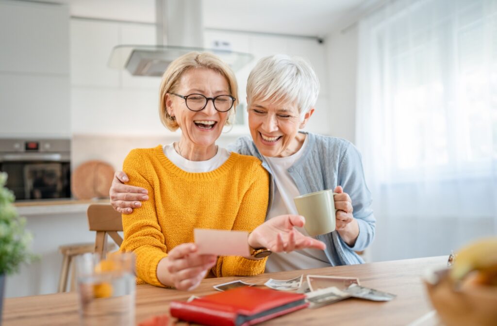Two senior women going through photos while seated at the table and exchanging broad smiles.