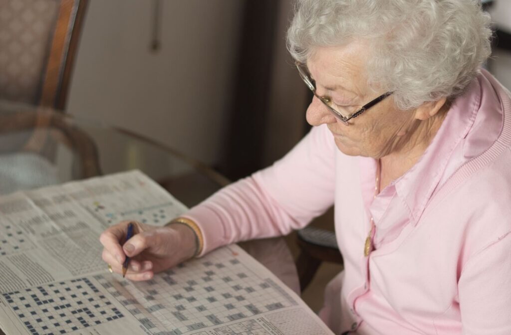 An older adult sitting at a table wearing glasses focuses on a crossword puzzle