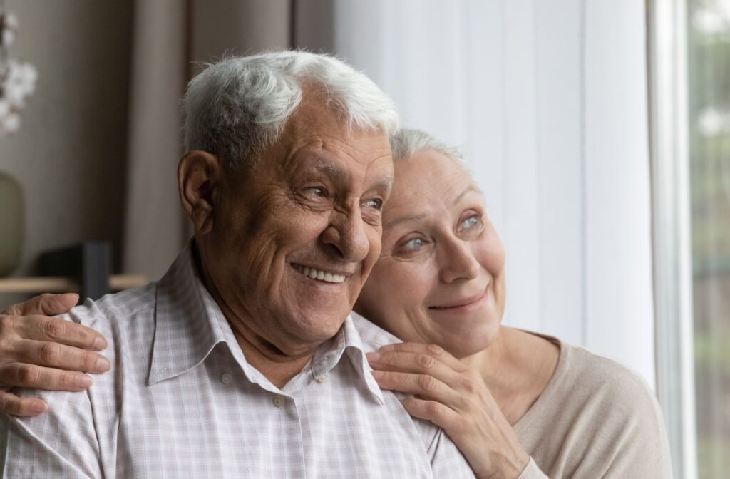 A smiling senior couple with Alzheimer’s sitting close together by a window in a calm, home-like memory care environment