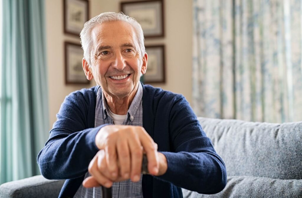 A smiling senior with Alzheimer’s is sitting on a couch and resting his hands on a cane in a comfortable memory care setting.