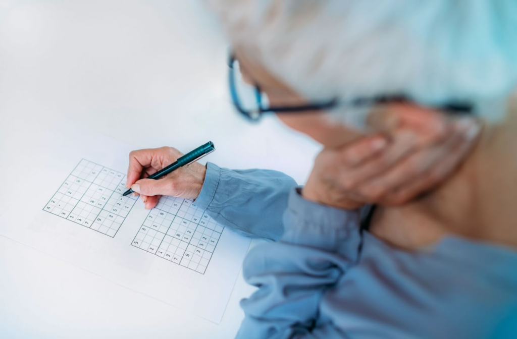 A senior in a blue shirt holds a pen above a square in a Sudoku puzzle, working on it to help with their memory
