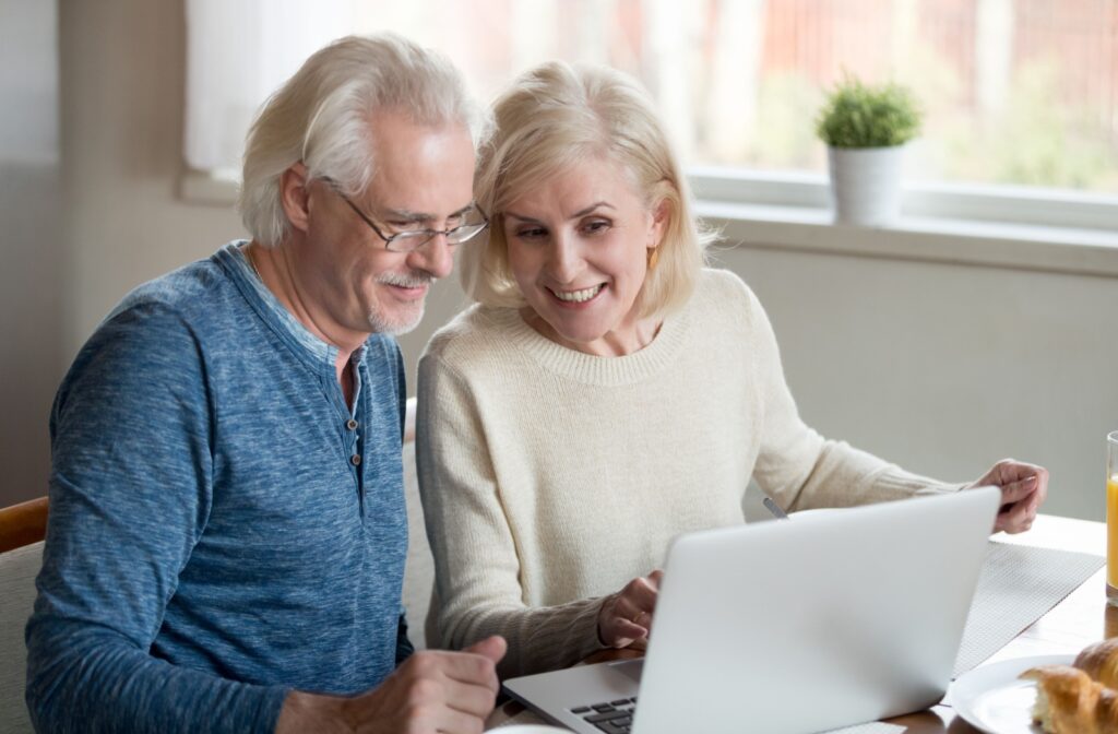 Two older adults sit at their table and smile while researching senior living communities on their laptop